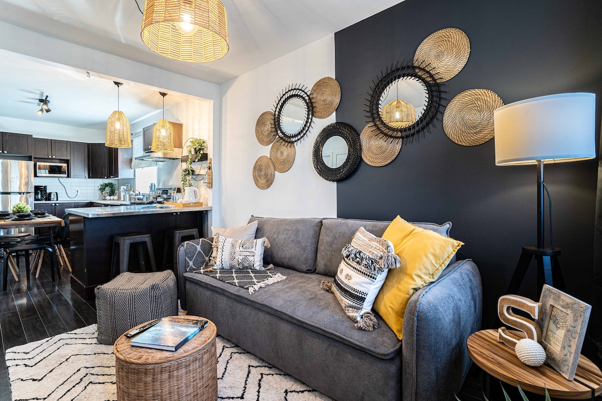 A cozy living area features a comfortable sofa adorned with decorative pillows. Surrounding wall decor includes various mirrors and woven baskets. Warm pendant lights hang above the open kitchen, which showcases modern appliances and a dining area, providing a welcoming atmosphere.