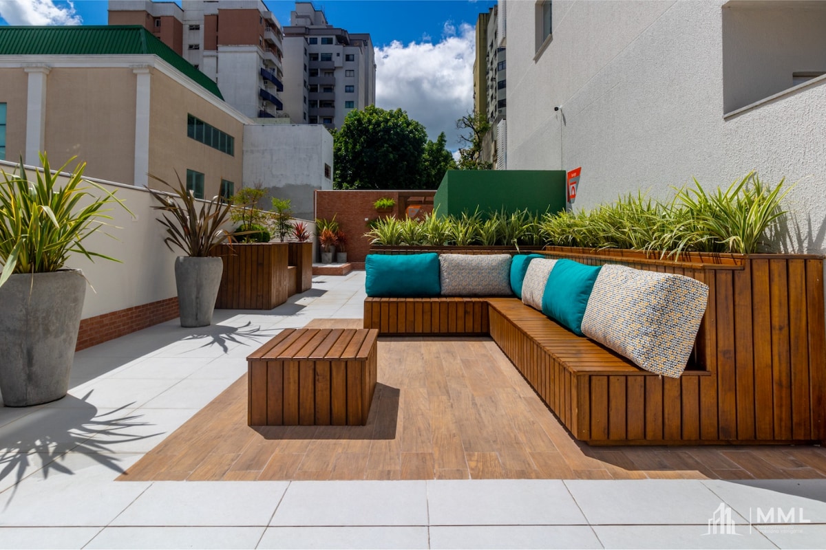 A spacious outdoor lounging area is presented with wooden seating, decorated with colorful cushions. Potted plants add greenery to the space, while a wooden coffee table complements the seating arrangement. The setting is designed for comfort and relaxation, surrounded by clean, modern architecture.
