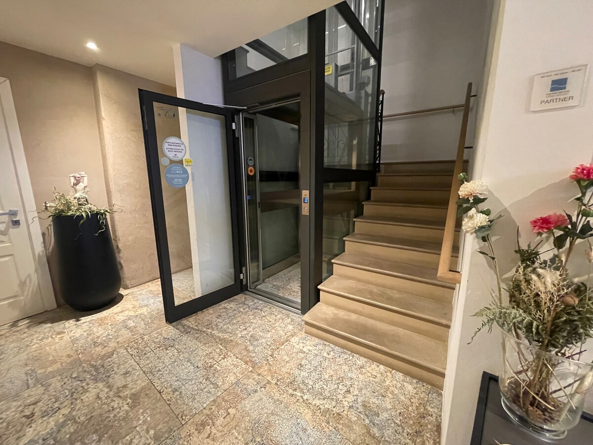 A welcoming entrance area is highlighted by a modern elevator and a staircase leading to upper floors. Natural stone tiles cover the floor, complemented by a potted plant and a simple floral arrangement on a nearby table.