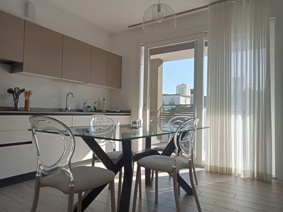 A modern kitchen area features a glass table supported by a black base, surrounded by transparent chairs. Light streams through large sliding doors leading to a balcony, with sheer white curtains providing a soft touch to the space.