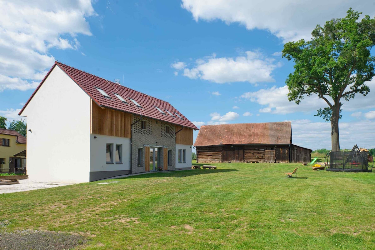 A spacious property is shown, featuring a modern two-story building with a combination of wood and brick elements. The large grassy area surrounds the structure, with a wooden barn visible nearby. The sky is partly cloudy, creating a bright and open atmosphere.