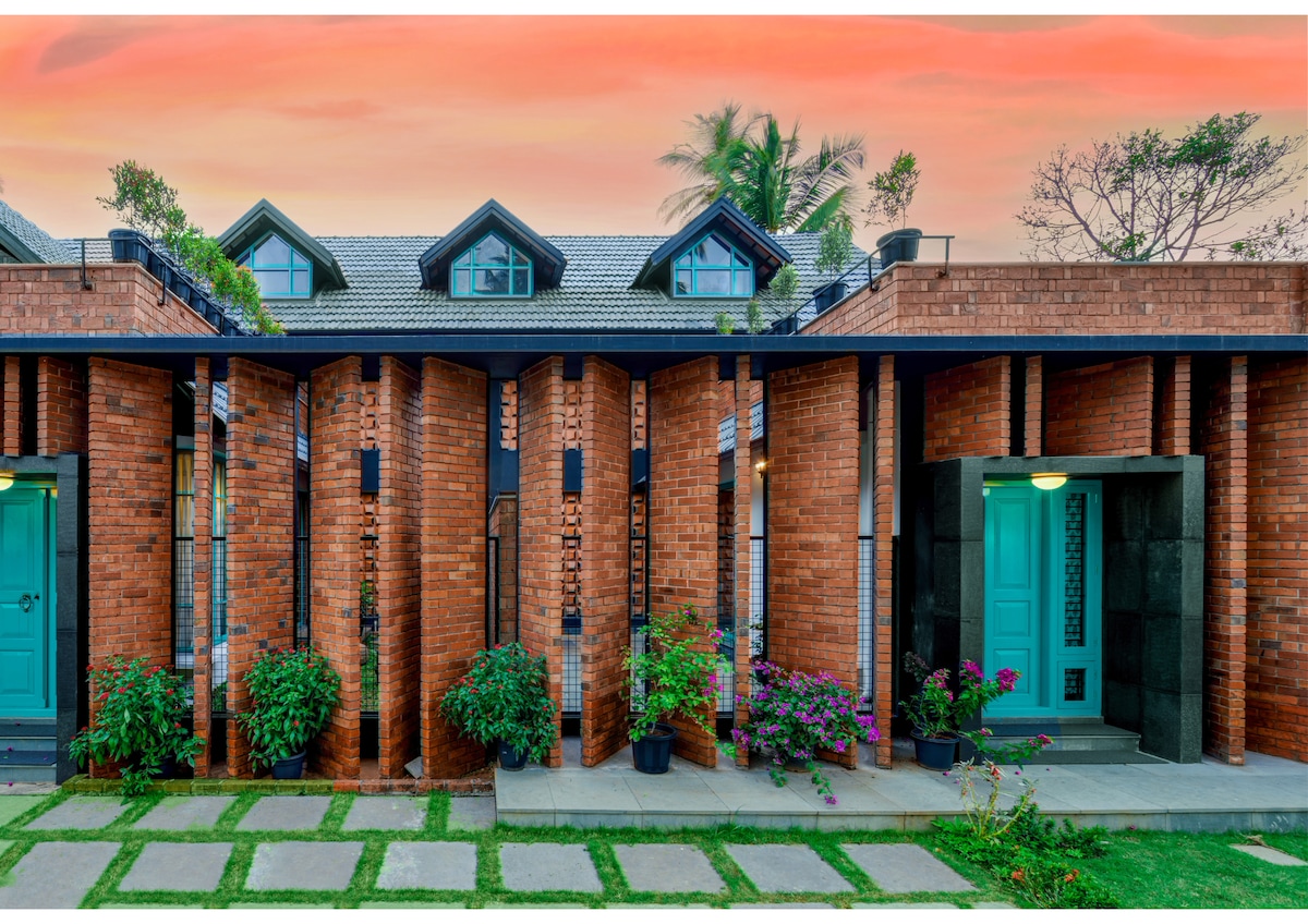 The exterior of the retreat is characterized by a modern design featuring brick walls and vibrant turquoise doors. Lush greenery and flowering plants enhance the entrance, while a well-maintained lawn and pathway lead to the doorways, creating a welcoming appearance.