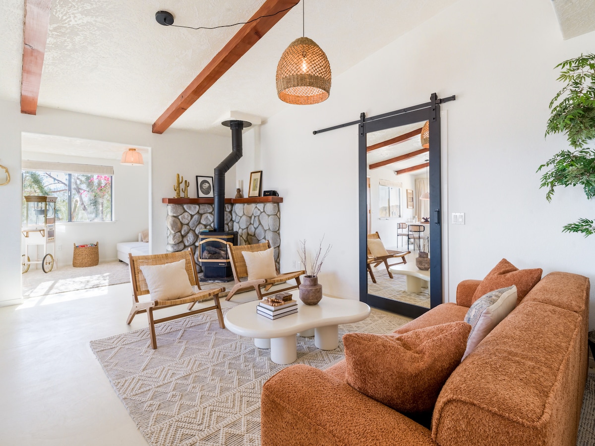 A cozy living area is highlighted by a modern sofa in a warm hue and natural fiber rug. A wood-burning stove is positioned against a stone feature wall. Large windows allow natural light to illuminate the space, featuring minimalist decor elements and an inviting atmosphere.