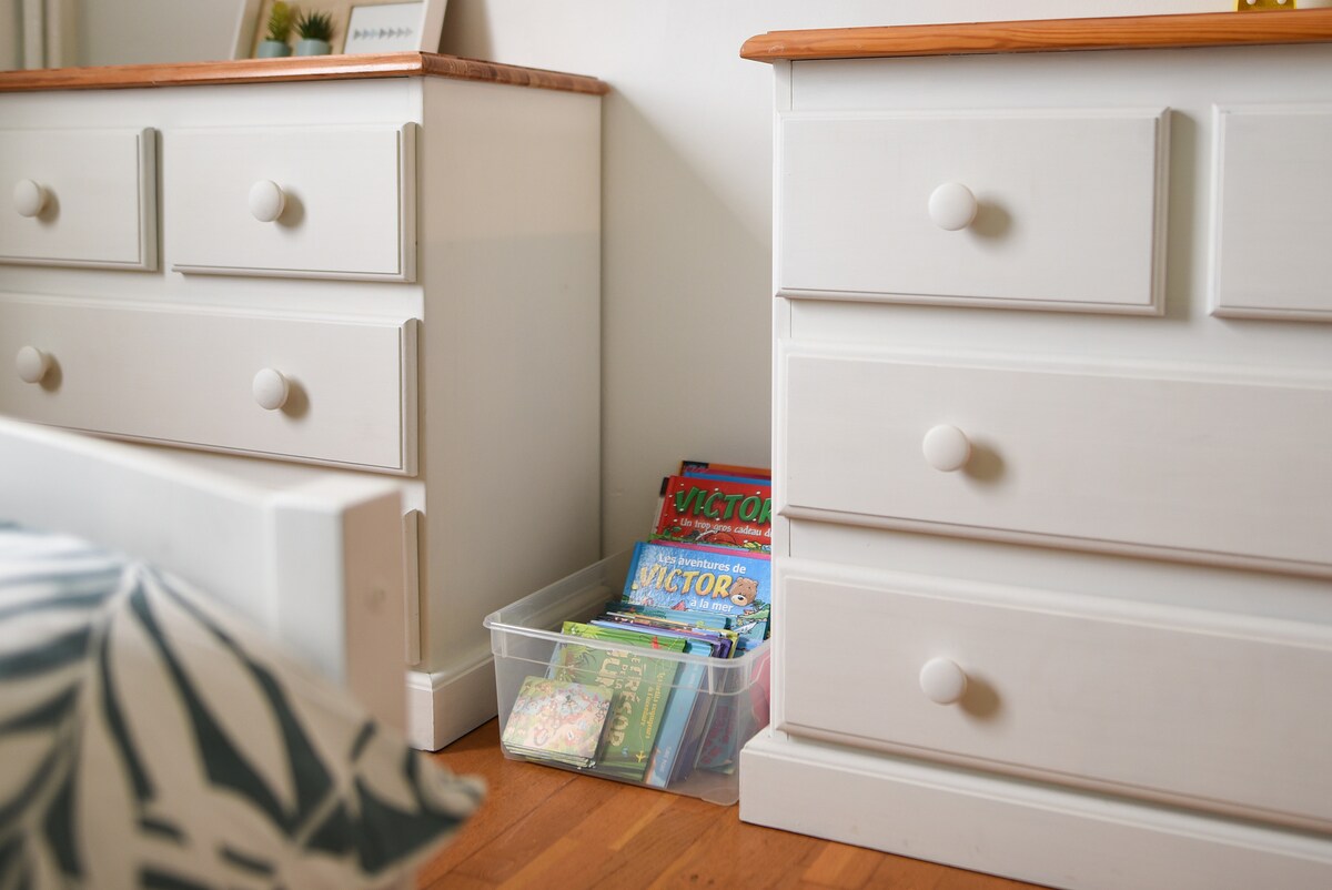 Two white dressers with round knobs are positioned beside a bed. A clear storage box filled with an assortment of children's books is placed on the floor between the dressers. Natural light softly illuminates the warm wooden flooring.