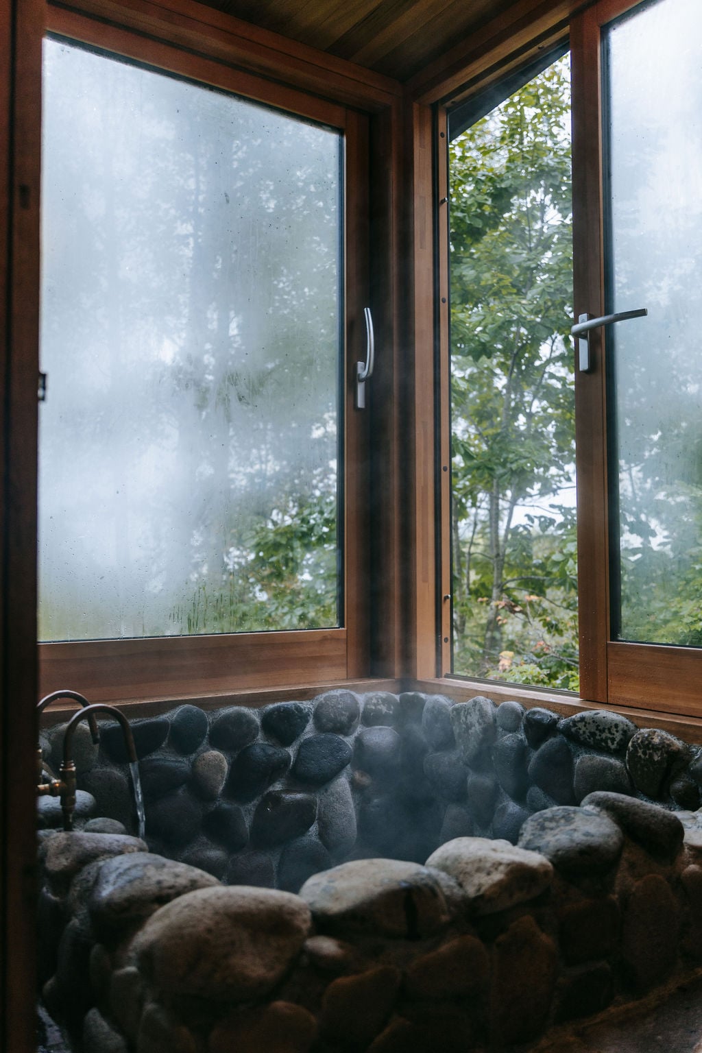 A traditional stone onsen is showcased, surrounded by large wooden-framed windows. Steam rises gently from the water, with views of the forest visible through the glass. The stone wall features rounded river stones, providing a natural and rustic aesthetic.