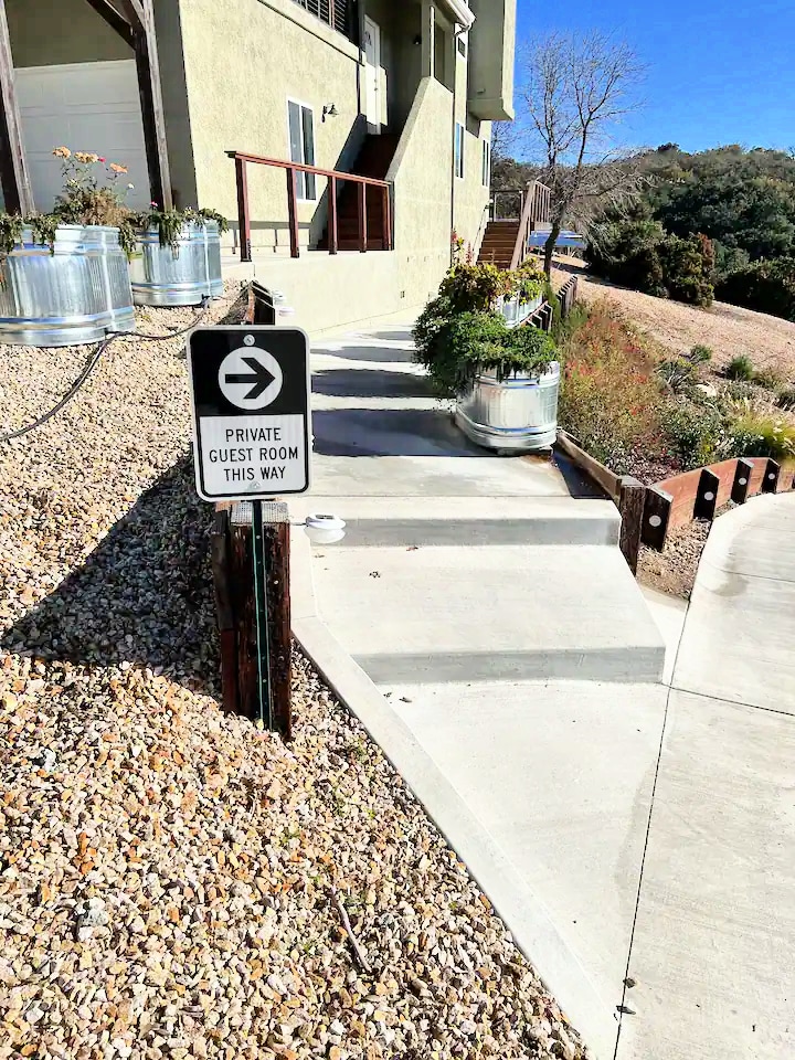 A pathway leading to the private guest room is shown, with a directional sign indicating the way. Potted plants are placed along the walkway, and a gravel area complements the concrete steps that guide guests. The surrounding landscape includes hills and trees.