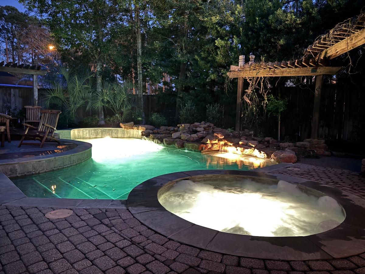 A private saltwater heated pool and hot tub are highlighted in soft illumination, nestled against a backdrop of lush greenery. Natural stone accents and a wooden pergola add an organic touch, while a paved area provides ample space for seating and relaxation.