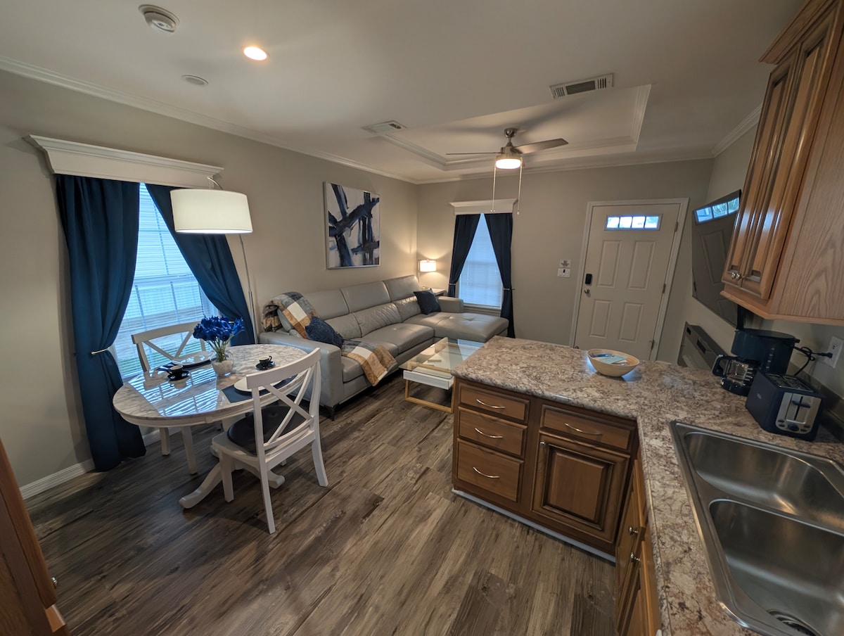 A cozy living area features a light gray sectional sofa paired with a glass-top coffee table. Natural light enters through the windows dressed in navy blue curtains. A dining table with white chairs and a decorative centerpiece is situated next to a well-equipped kitchen with wood cabinets and modern appliances.