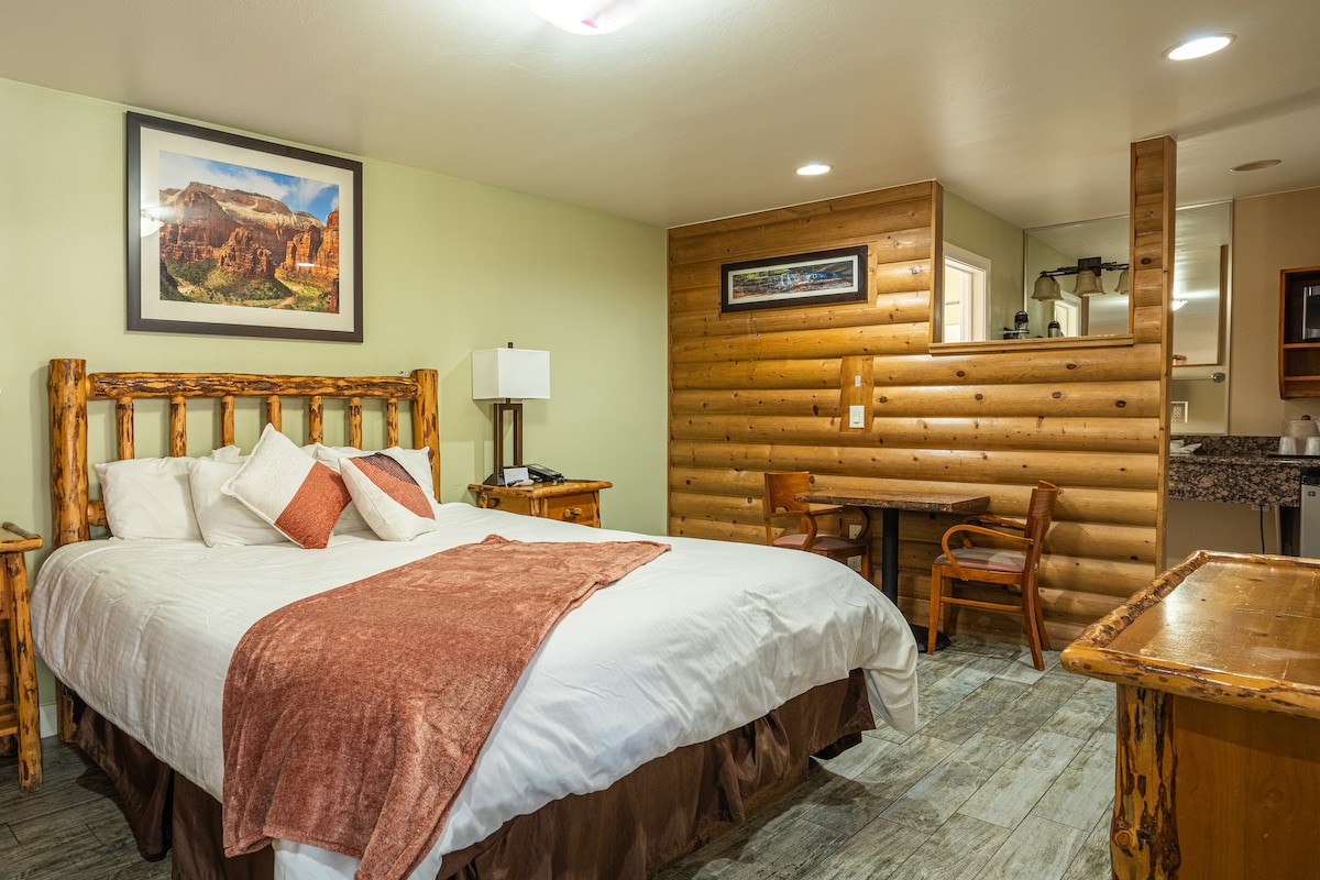 A spacious room features a King-size bed with a warm brown blanket and decorative pillows. A rustic wood accent wall is visible behind the bed. A small dining table and chair are positioned against the side wall. A kitchenette area is also included.