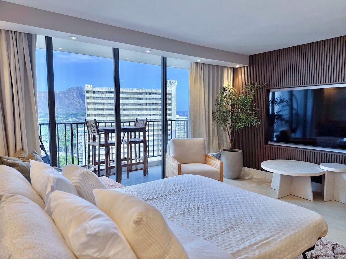 The spacious living area features a comfortable queen pull-out sofa and a large modular sectional, positioned near floor-to-ceiling windows that offer stunning views of Diamond Head. A 75-inch HDTV is mounted on the wall, and a small dining setup is visible on the balcony.
