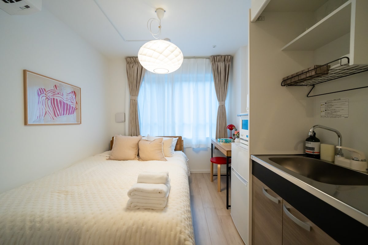 A compact room features a spacious bed dressed in light bedding, two soft pillows placed at the head. A window with sheer curtains allows natural light to brighten the space. A small dining table with a red chair and a kitchenette with essential appliances complete the room's layout.