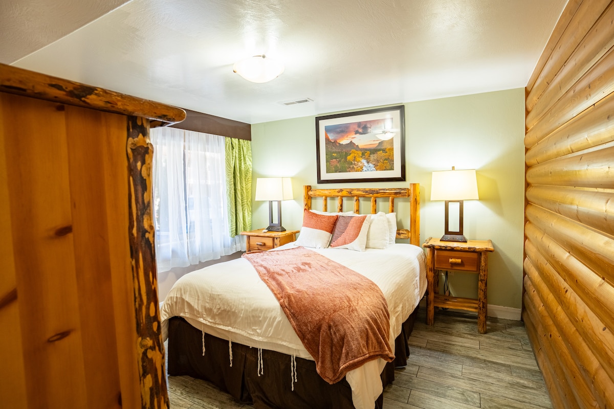 A cozy bedroom features a spacious bed with a light-colored blanket and decorative pillows. Two bedside tables with lamps are positioned on either side of the bed. A piece of artwork hangs above the headboard, while light filters through the curtains, illuminating the rustic wood accents.