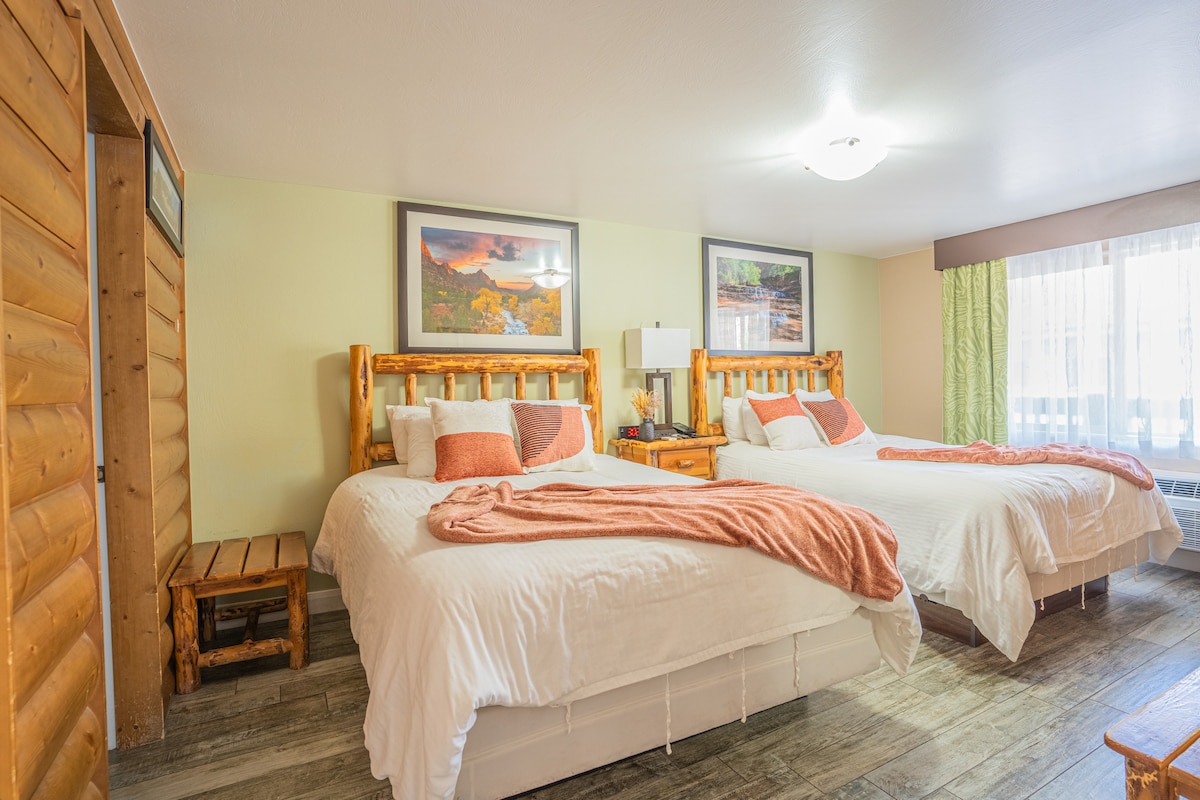 A spacious room features two Queen-size beds with warm bedding, set against a backdrop of wooden accents. Natural light flows through a large window, complemented by sheer and room-darkening curtains. A cozy seating area with a wooden bench is positioned nearby for relaxation.