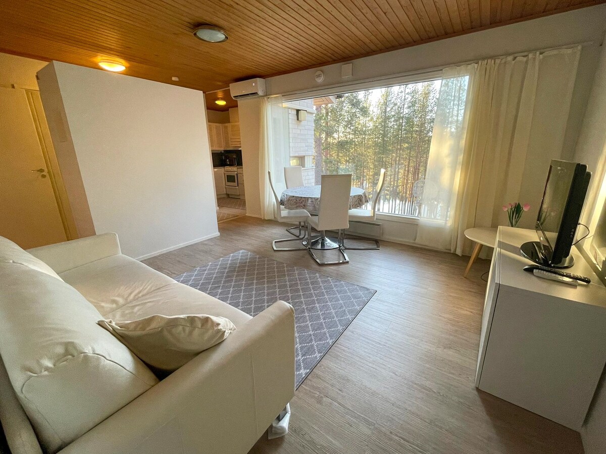 A light-filled living area is captured, featuring a white sofa and a round dining table set for four. Large windows allow for views of surrounding trees, with sheer curtains softening the natural light. A minimalist TV unit stands opposite the sofa.