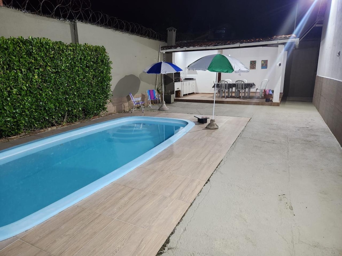 A private outdoor area features a rectangular pool, surrounded by tiled decking. Two umbrellas provide shade over a set of outdoor tables and chairs. Lush green hedges line one side, enhancing the spacious layout under night lighting.