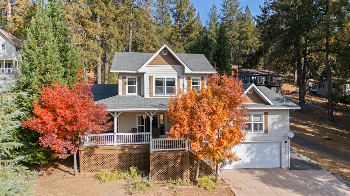 The exterior of the home is presented, showcasing a two-story structure surrounded by trees. A covered porch is visible, along with a garage. Vibrant red and orange foliage from nearby trees adds color to the setting, complemented by a clear blue sky.