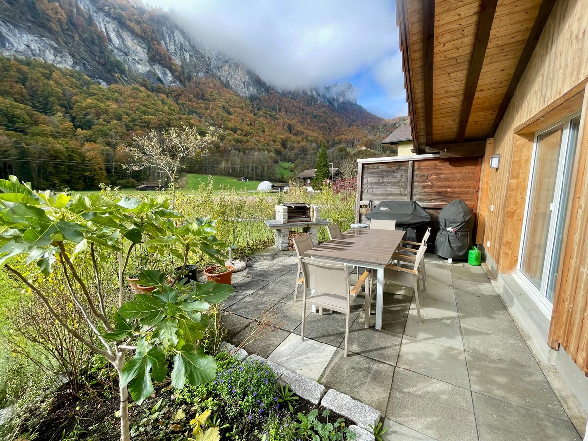 An outdoor patio area is shown, featuring a wooden table with six chairs surrounded by lush greenery and decorative plants. A BBQ grill is visible next to a wooden shed. The backdrop includes a mountainous landscape with autumn foliage under a partly cloudy sky.