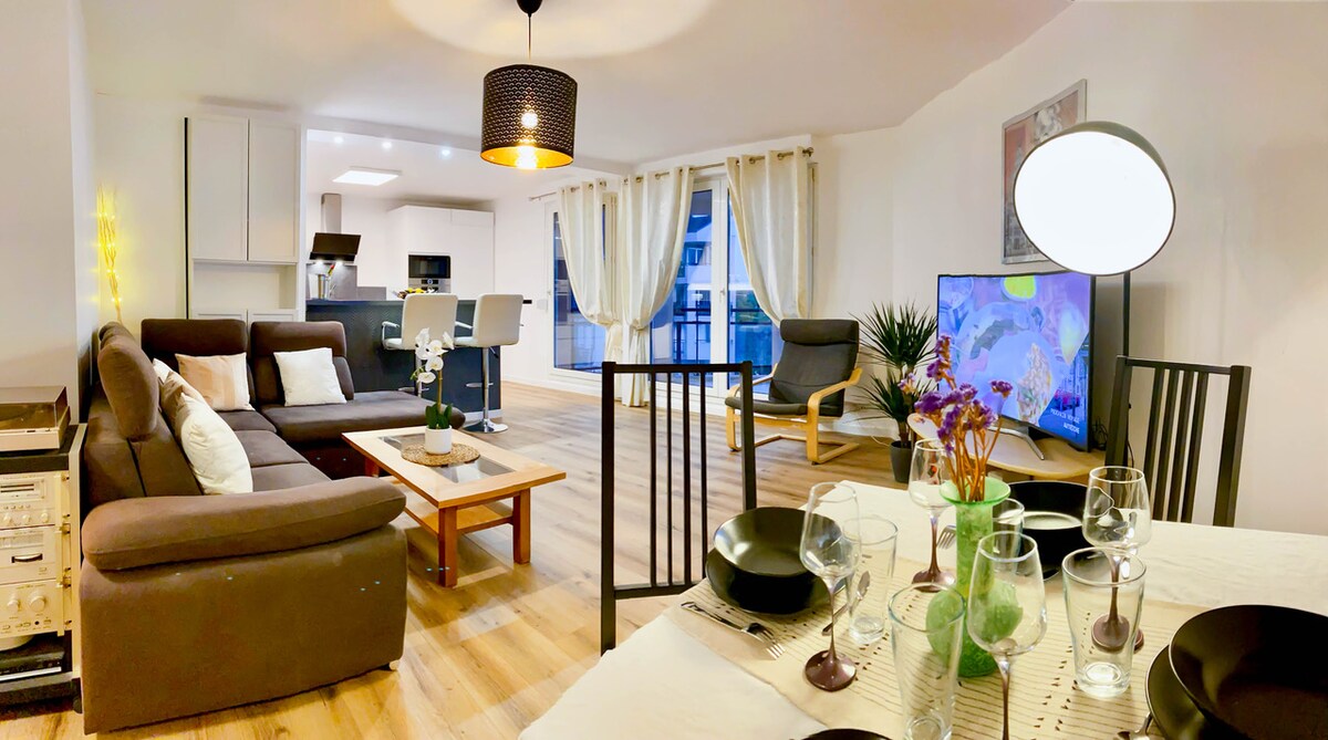 An open living area features a comfortable seating arrangement with a brown sofa and two accent chairs. A dining table set for six is present, accompanied by decorative greenery. A large TV displays content, while balanced natural light filters through sheer curtains, enhancing the space.