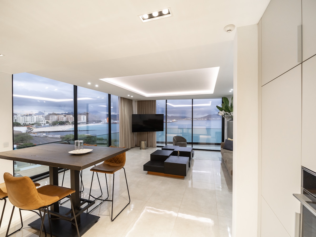 A contemporary living space showcases floor-to-ceiling windows that provide panoramic views of the coastline. A dark wood dining table is accompanied by four high-backed chairs, while a minimalist black couch and a sleek television add to the modern aesthetic. Natural light enhances the open layout.