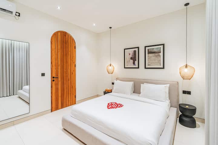 Una camera da letto ordinatamente disposta con un grande letto al centro. Il letto ha lenzuola bianche e due cuscini, con una disposizione a forma di cuore di petali rossi in cima. Ci sono due sospensioni a sospensione, un comò in legno e un alto armadietto in legno.