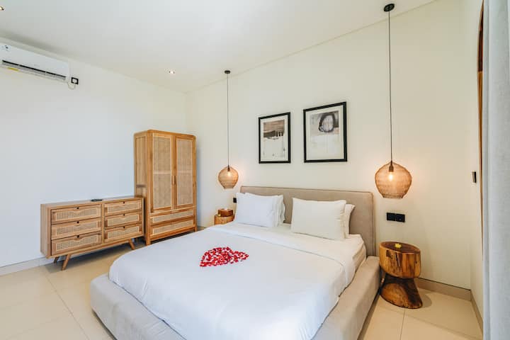 Una camera da letto ordinatamente disposta con un grande letto al centro. Il letto ha lenzuola bianche e due cuscini, con una disposizione a forma di cuore di petali rossi in cima. Ci sono due sospensioni a sospensione, un comò in legno e un alto armadietto in legno.