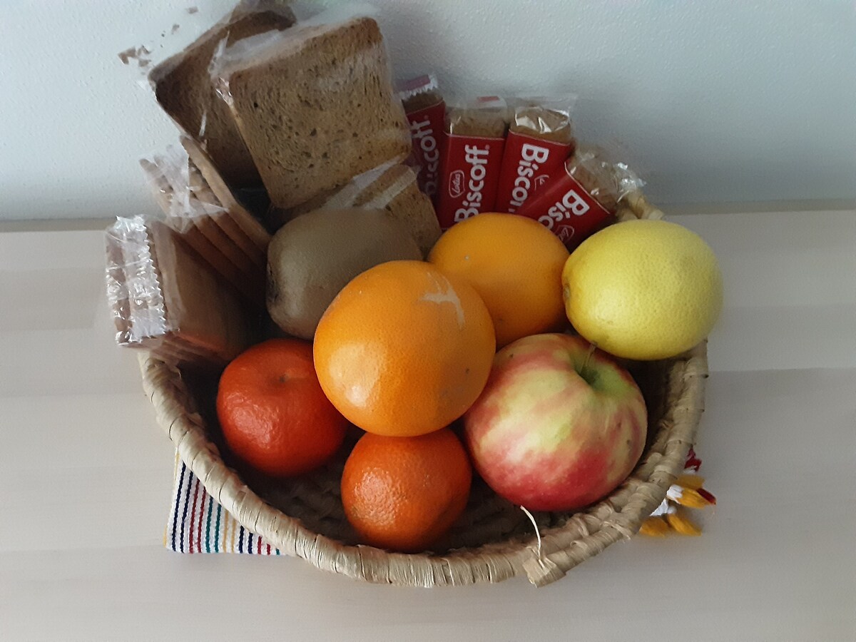A woven basket contains a variety of items, including several types of fruit such as apples, oranges, and lemons, alongside slices of brown bread and packaged biscuits. The arrangement creates an inviting presentation for guests.