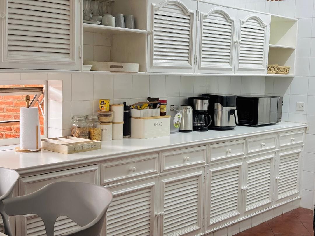 A well-organized kitchen features white cabinetry with louvered doors and a countertop that accommodates various kitchen appliances, including a coffee maker and microwave. Glass jars filled with ingredients are visible, and a small window provides natural light, enhancing the bright ambiance.