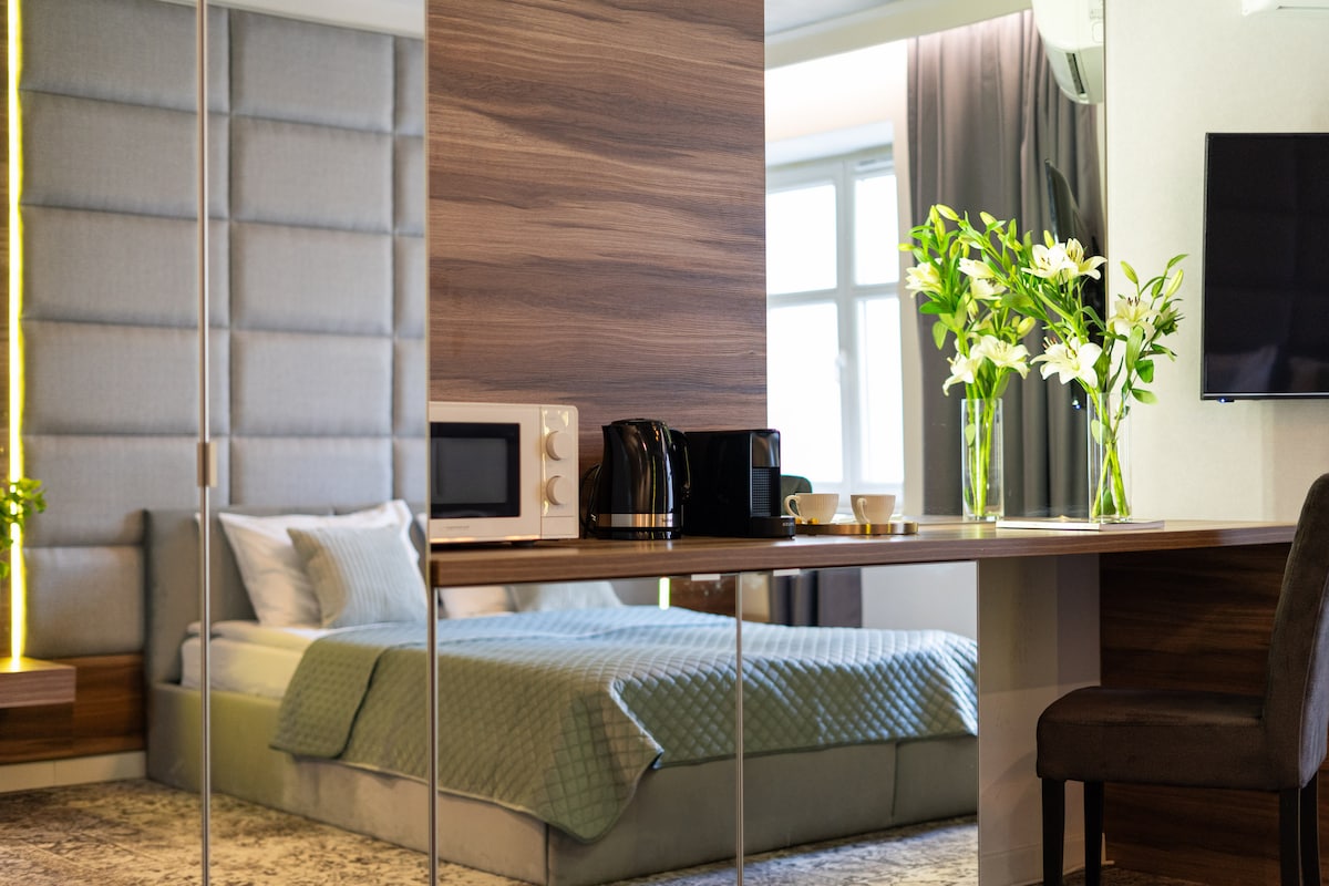 A well-appointed space features a spacious bed and a kitchenette area with a microwave and coffee maker. A small table is set with a flower arrangement, adding a touch of nature. The room is brightened by natural light filtering through the window.
