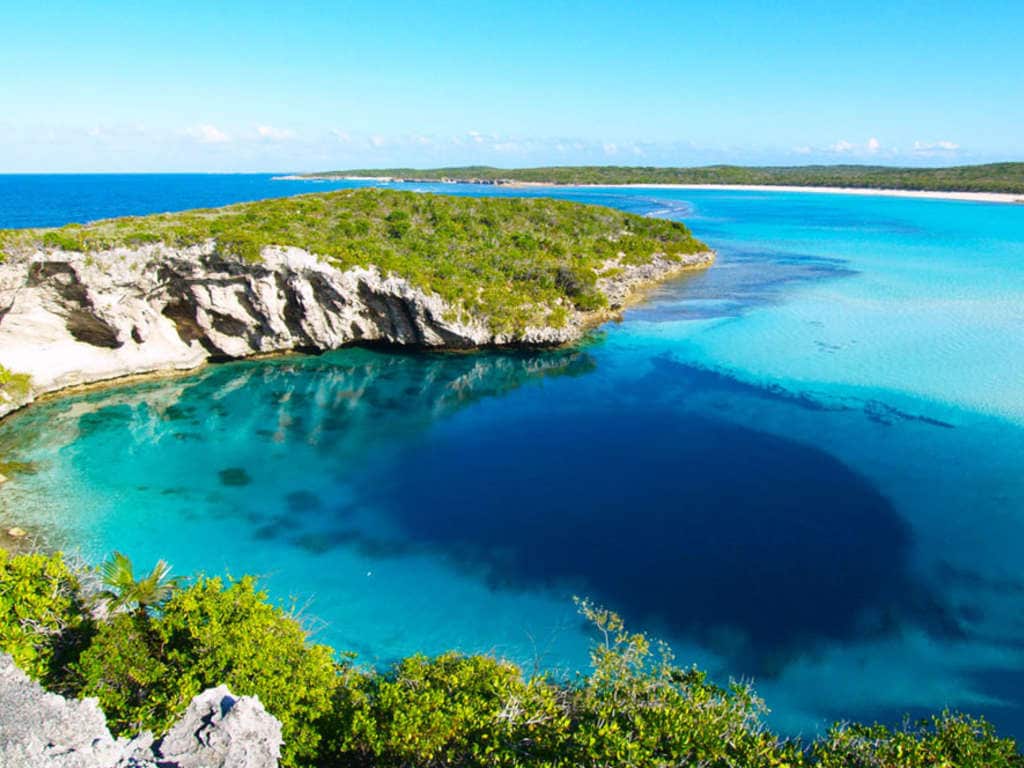 A serene coastal landscape displays a vibrant blue lagoon surrounded by lush greenery. Gentle waves lap against rocky shorelines, while sandy beaches are visible in the distance, merging with the deep blue of the ocean under a clear sky.