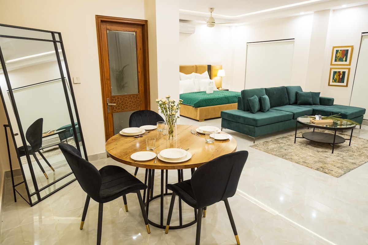 A spacious living area features a comfortable L-shaped green sofa and a round wooden dining table set for four. A large mirror reflects the contemporary decor, and soft lighting accentuates the elegant design. The inviting atmosphere is complemented by tasteful wall art.