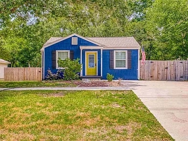A charming blue cottage is nestled in a leafy setting, featuring an inviting yellow door as a focal point. Flanking windows are framed by dark shutters. The well-kept front yard is bordered by a wooden fence, while a driveway provides ample parking space.