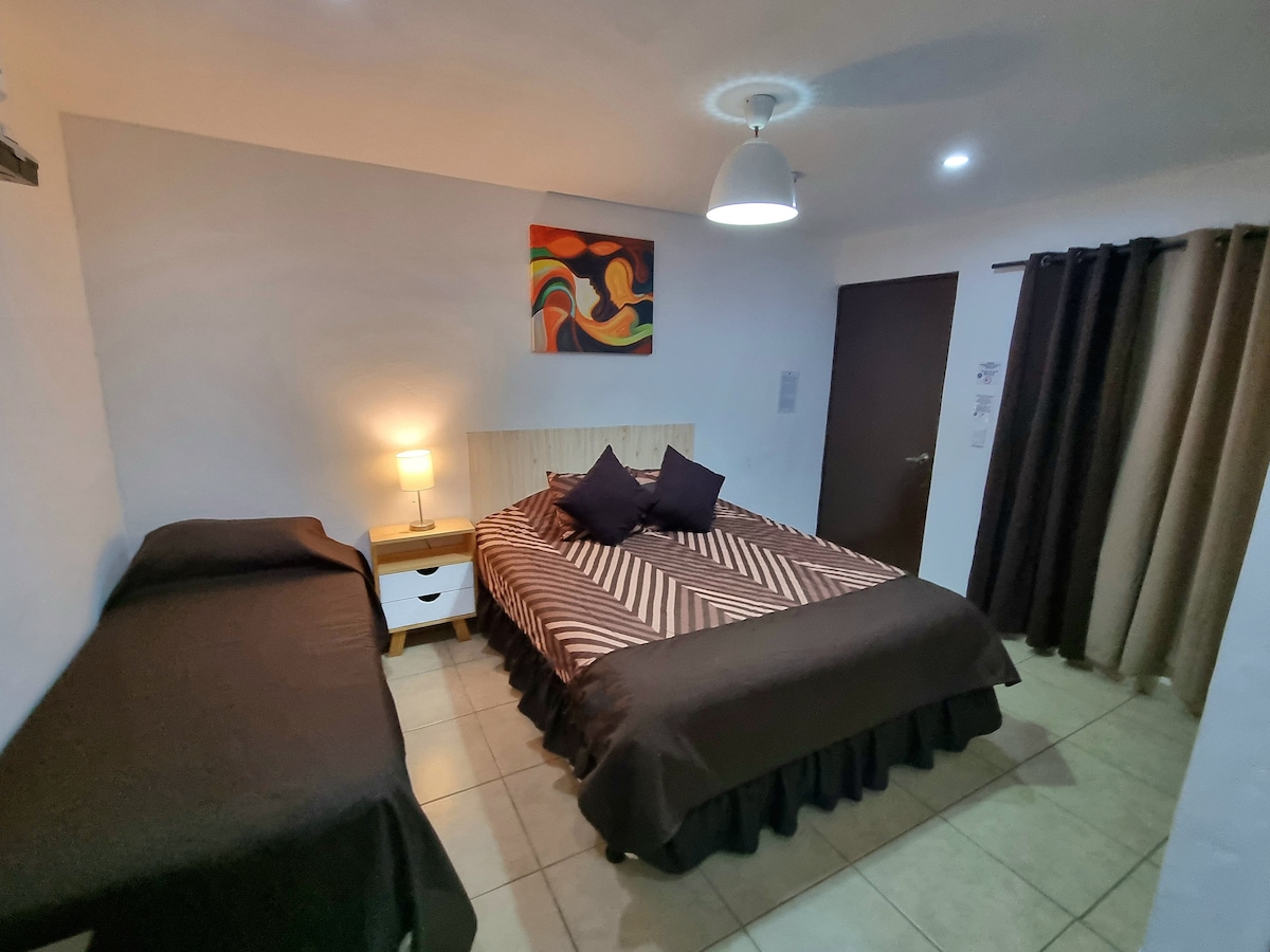 A cozy room features a spacious bed with a patterned duvet and two accent pillows. A single bed is positioned beside a bedside table with a lamp. A colorful piece of art hangs above the bed, and soft lighting is provided by an overhead fixture.