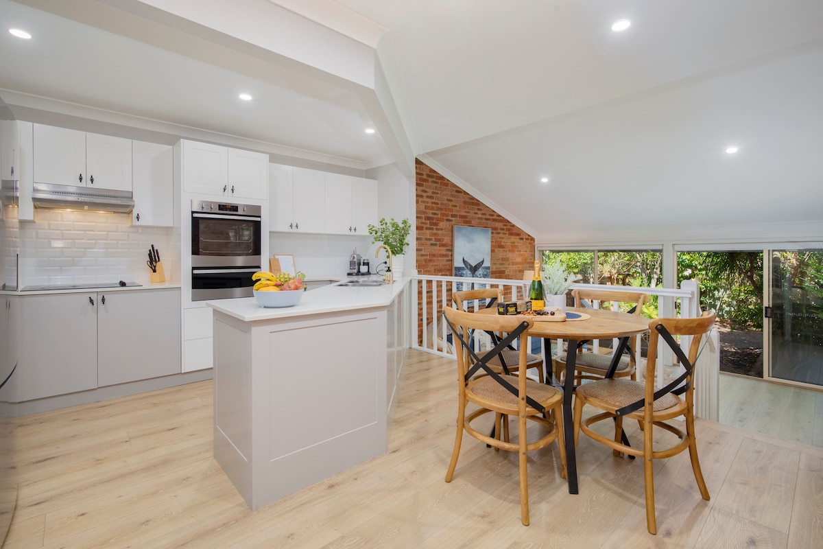 The modern kitchen features sleek white cabinetry and stainless steel appliances, with a spacious island at the center. A light-filled dining area is visible, furnished with a wooden table and chairs, offering a view of the garden beyond.