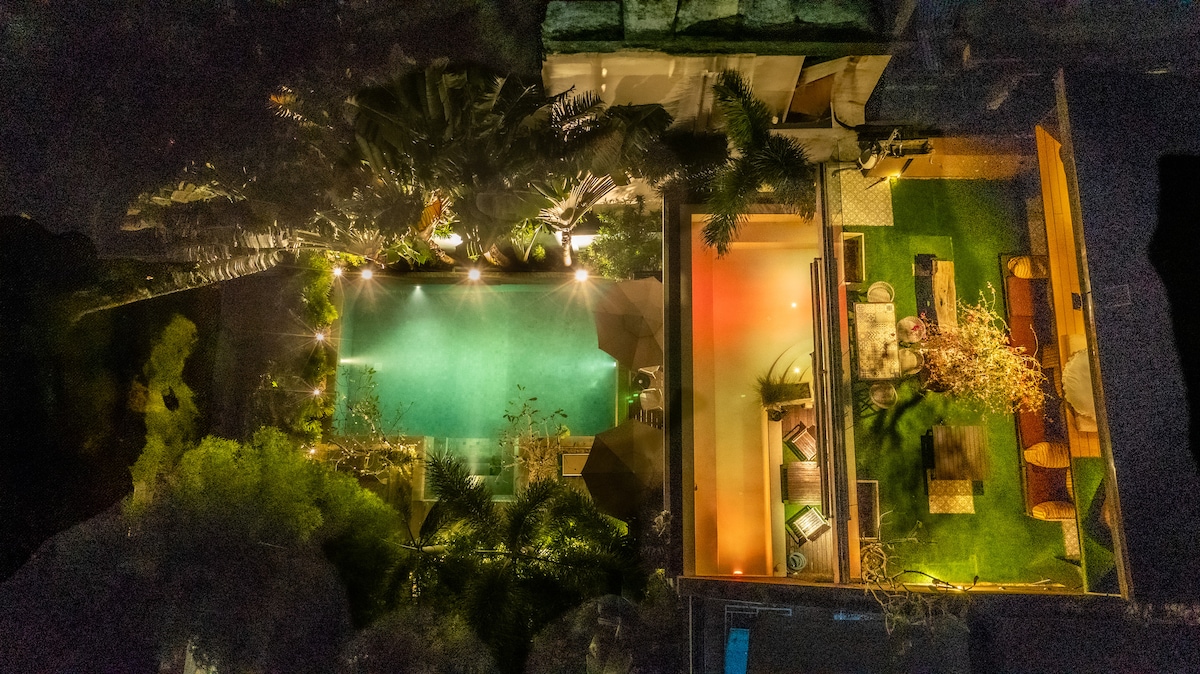 An aerial view showcases a spacious property featuring a dual swimming pool surrounded by greenery. One pool area is illuminated, while a nearby outdoor lounge on a grassy surface is visible, presenting a blend of relaxation and leisure options, all set within a tranquil environment.