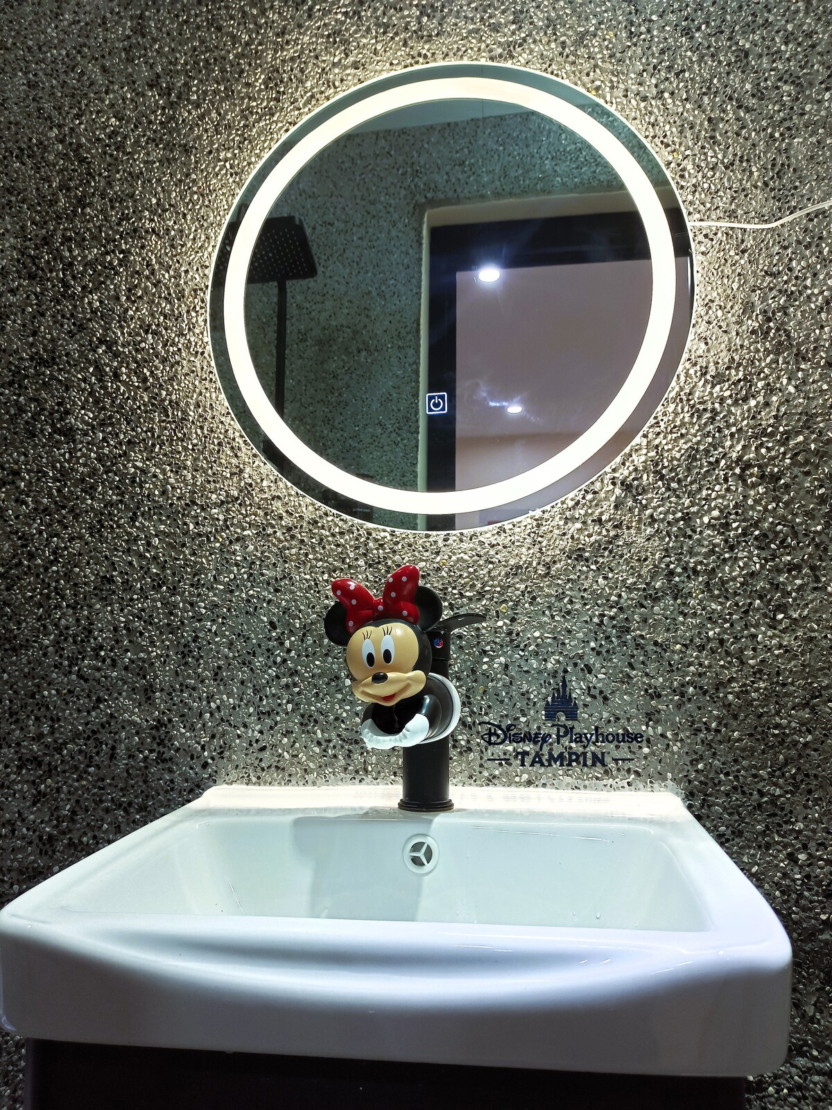 A modern bathroom sink is illuminated by a circular mirror featuring a soft glow. A whimsical faucet handle shaped like Minnie Mouse adds a playful touch to the sleek design. The textured wall serves as a contemporary backdrop, enhancing the overall aesthetic.