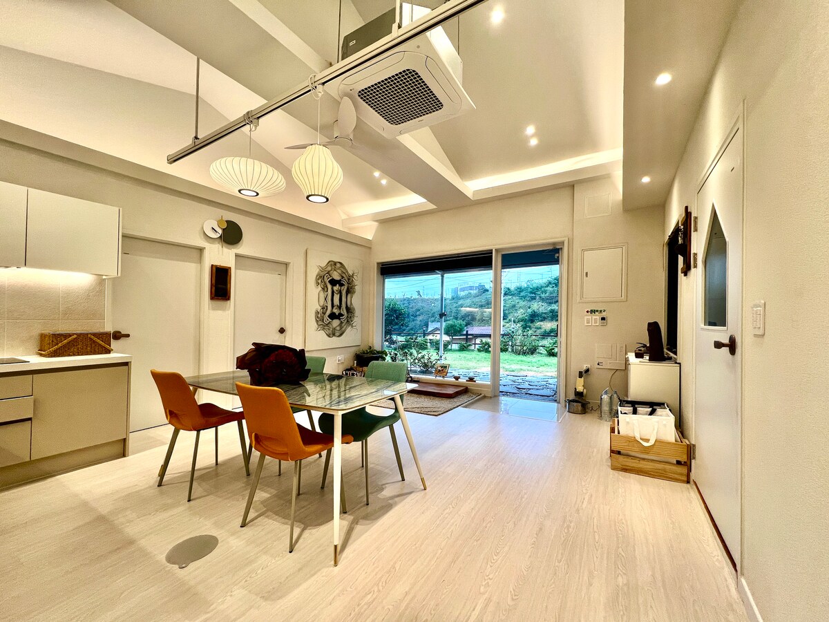 An open-concept living space is highlighted by modern furnishings, featuring a glass dining table surrounded by four colorful chairs. Floor-to-ceiling windows provide a view of the outdoors, while a cozy seating area is situated in the corner, creating a welcoming atmosphere.