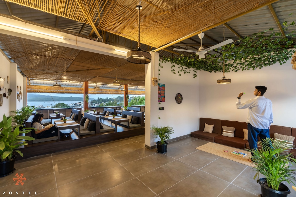 A spacious common room features a relaxed seating area with dark couches and low tables, surrounded by greenery and decorative plants. Large windows offer a view of the sea, while a ceiling fan provides ventilation. Warm lighting enhances the inviting ambiance of the space.