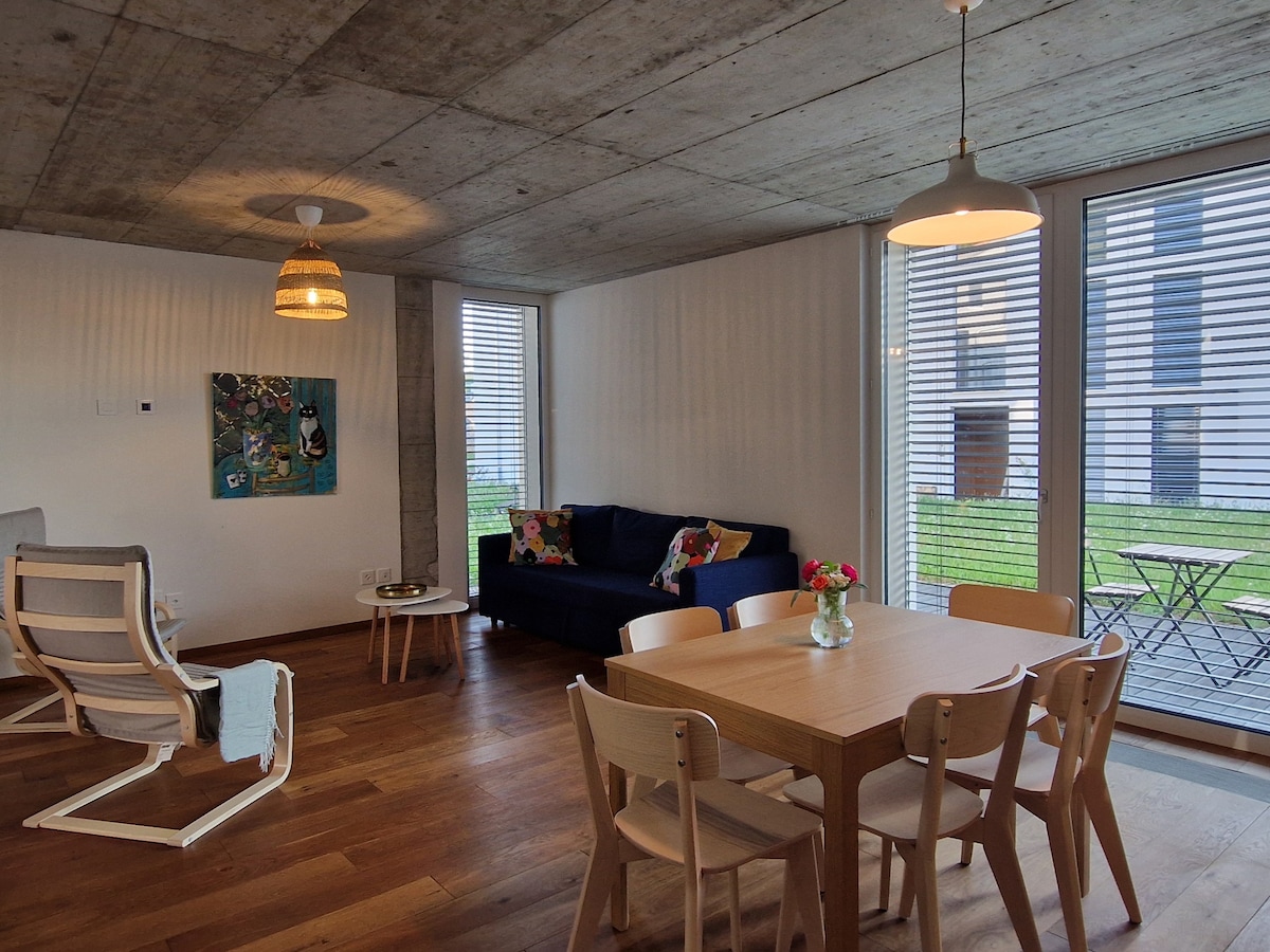 An open living space features a modern sofa with colorful cushions, a dining table, and light wood chairs. Natural light enters through large windows, illuminating the polished wooden floors. A small table holds a vase of flowers, enhancing the fresh atmosphere.