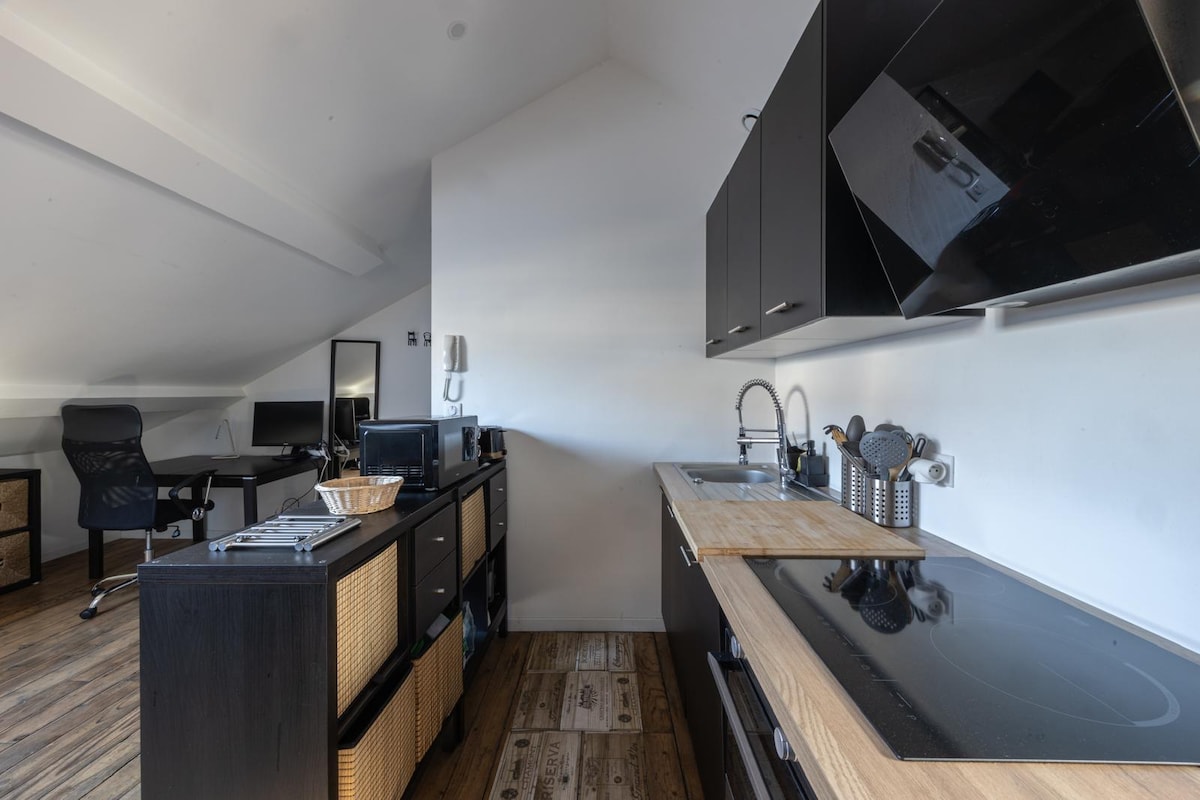 A modern kitchen space is equipped with sleek black cabinets and a light wooden countertop. Essential appliances are organized neatly, including a stovetop and sink. A small workspace area is seen in the background, enhancing the functionality of the room.