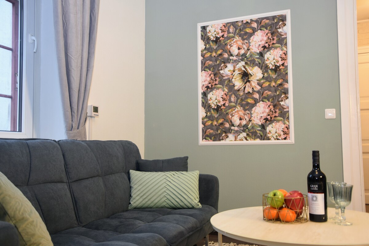 A cozy seating area presents a dark upholstered sofa, accented by a textured throw pillow. A light-colored coffee table sits in front, adorned with a bowl of fresh fruits and a bottle of wine. A floral wall art piece adds a touch of color to the room.