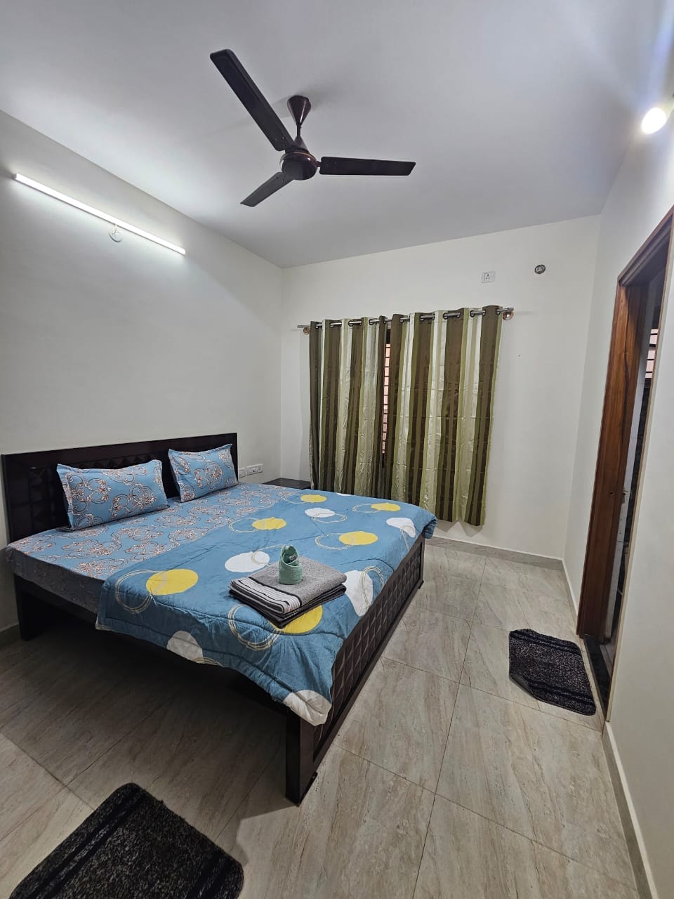 A well-appointed bedroom features a large bed dressed in a colorful bedding set adorned with patterns, complemented by two supportive pillows. A ceiling fan provides airflow, while light filters through two sheer curtain panels. A small plant is placed on the bed, adding a touch of greenery.