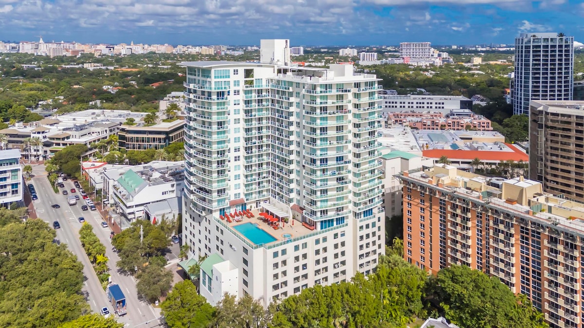 An aerial view captures a modern high-rise building surrounded by lush greenery and urban developments. The rooftop pool area is visible, featuring lounge chairs and views of the cityscape in the background, showcasing a blend of nature and city life.