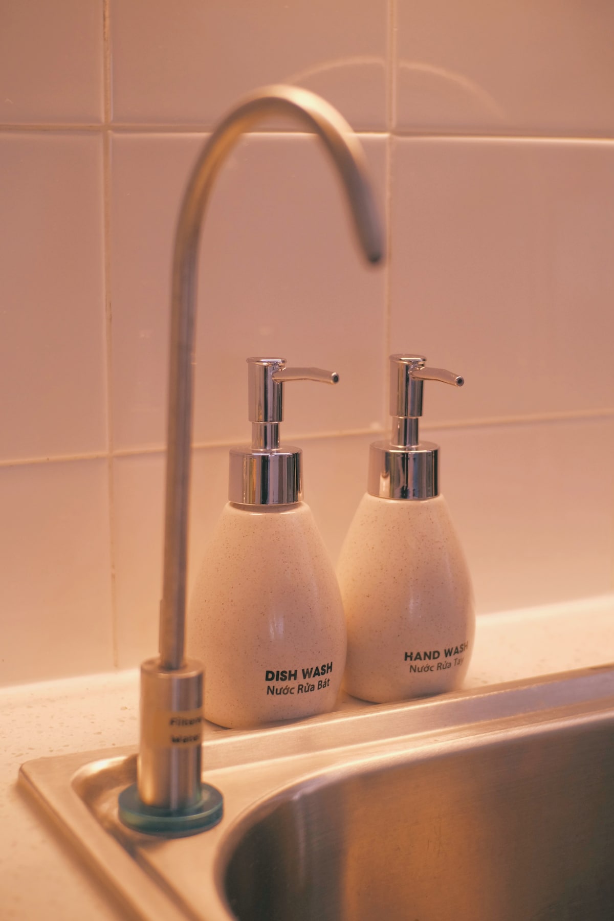 Two sleek dispensers for dish wash and hand wash are positioned beside a modern kitchen faucet. The minimalist design complements the clean, tiled background, creating a functional and tidy kitchen space.