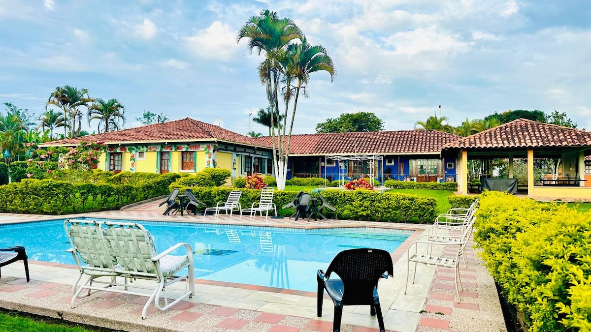 A serene pool area is surrounded by colorful landscaping and several palm trees. The inviting swimming pool reflects the sky, while comfortable lounge chairs are placed around it. The rustic building features a tile roof and multiple entrances, presenting a harmonious blend with the natural environment.