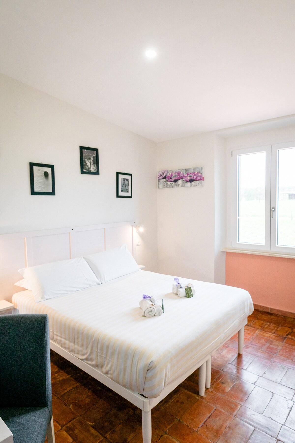 The room features a spacious bed with white linens and decorative pillows arranged neatly. Artwork decorates the walls, complemented by a small window allowing natural light to fill the space. A cozy seating area is visible, enhancing the comfortable atmosphere.