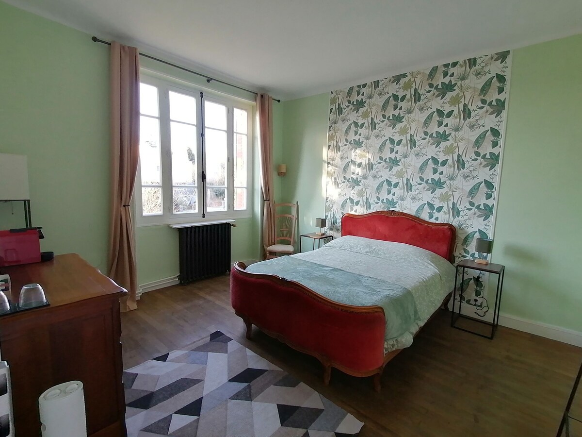 A double bedroom features a spacious bed with a gently curved red headboard and soft green bedding. Natural light filters through large windows, complemented by light drapes. A patterned accent wall adds a touch of color, while a wooden dresser and simple chair complete the inviting space.