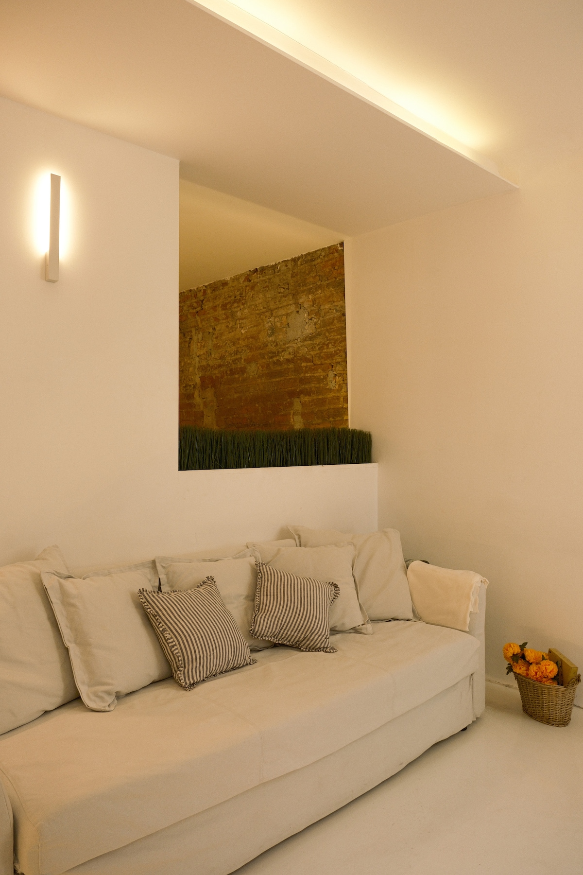A neutral-toned living area features a light-colored sofa adorned with various patterned cushions. A brick wall partially reveals greenery above, creating an inviting backdrop. Soft lighting enhances the cozy setting.