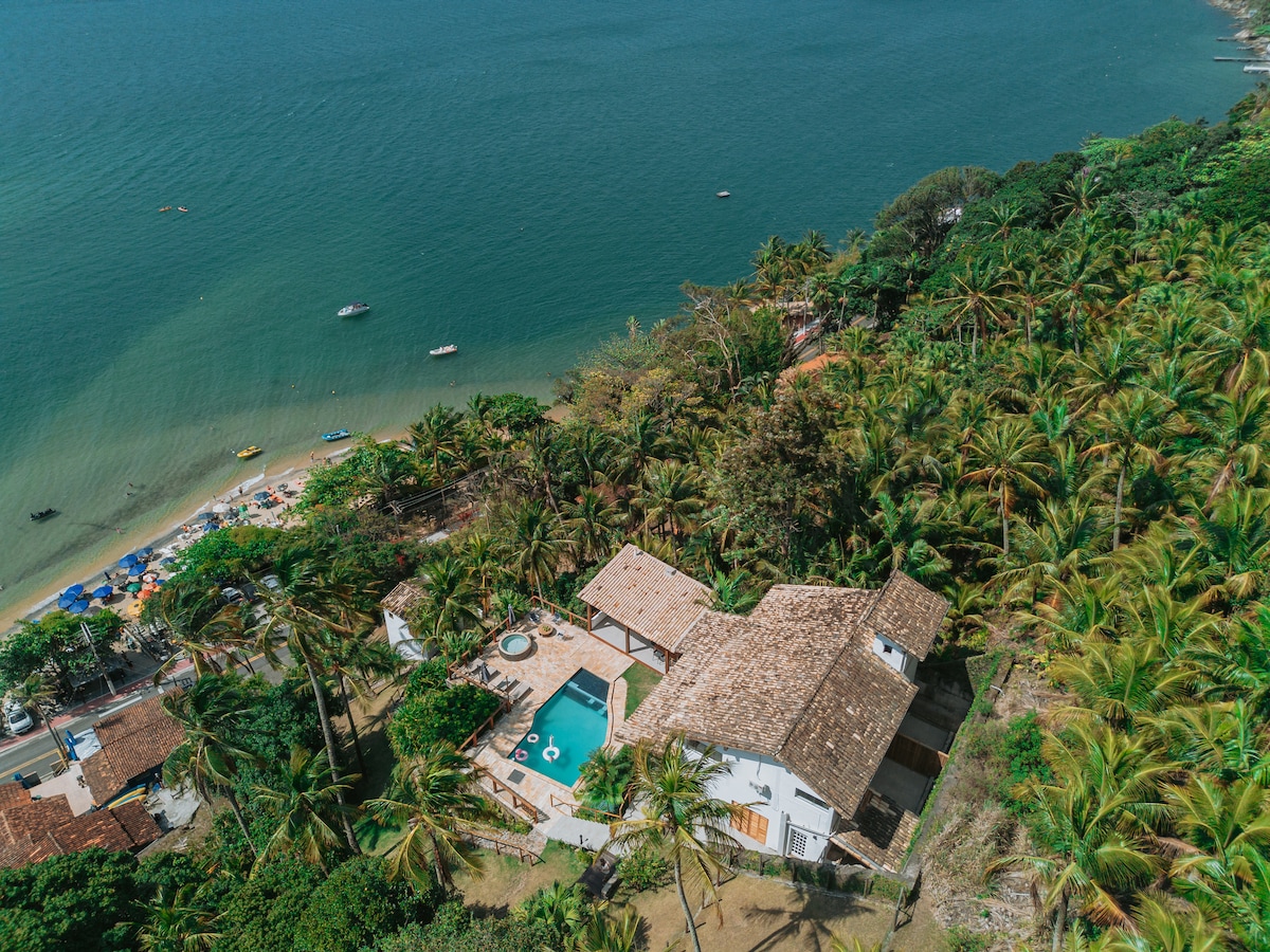 An aerial view of a spacious house nestled among lush palm trees near the beach. A clear blue pool is visible in the backyard, surrounded by sun loungers. The coastline, with its sandy beach and boats, is seen in the background, highlighting the property's scenic location.