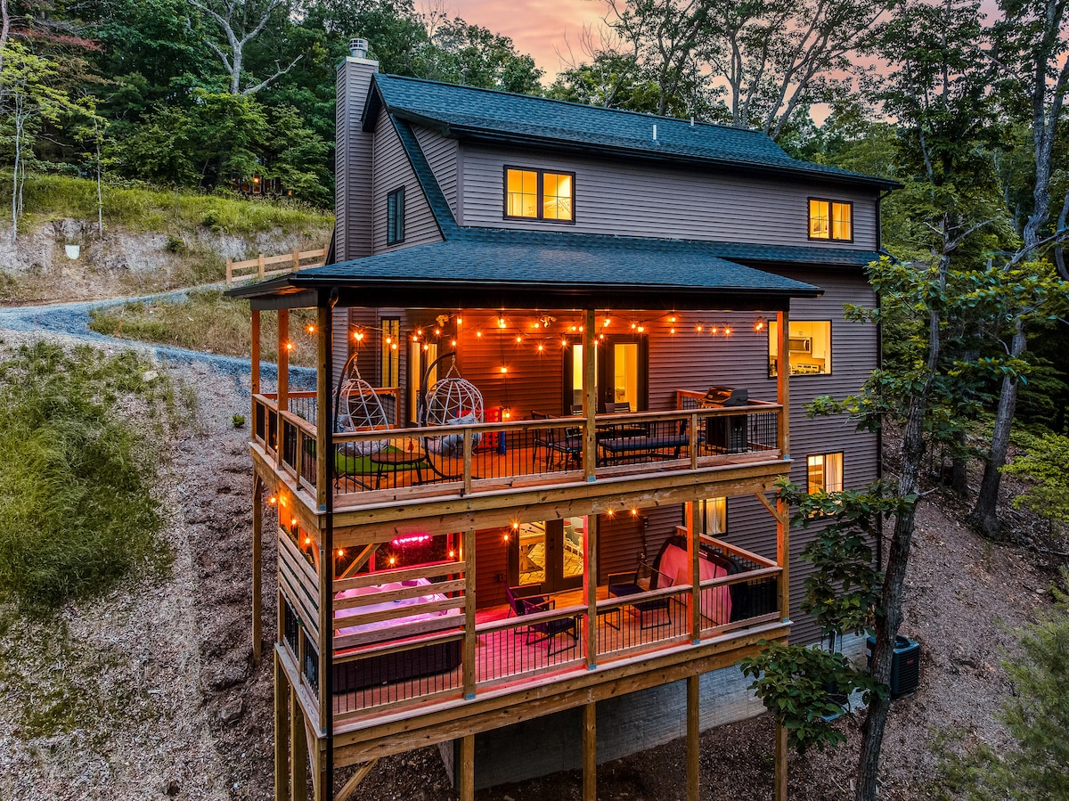 A modern, three-level home is framed by trees, featuring multiple lighted balconies and a deck. String lights adorn the outdoor space, creating a welcoming ambiance. Large windows reflect the surrounding nature, inviting natural light into the living spaces.