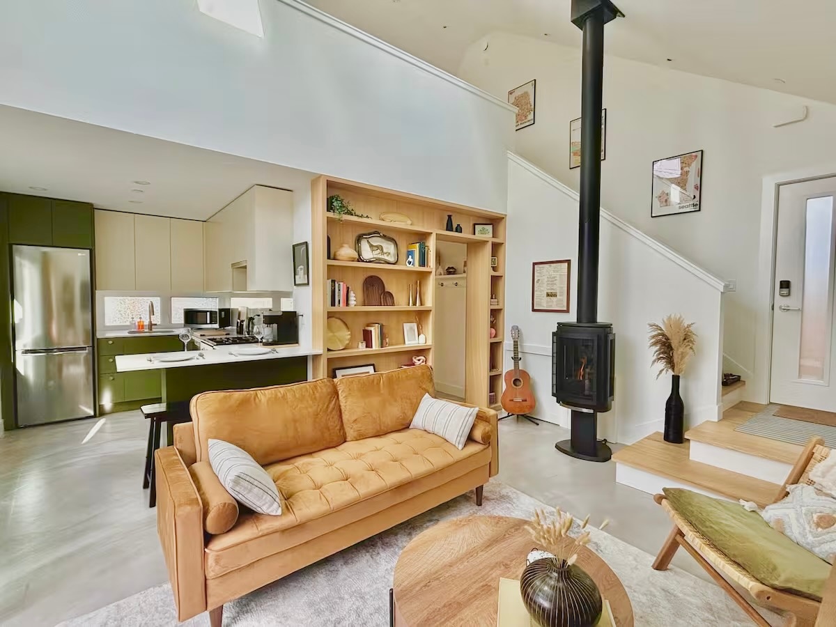An open living space showcases a cozy brown sofa with decorative pillows, next to a round coffee table. A modern kitchen with sleek finishes is visible in the background, highlighted by wooden shelves displaying books and decor. A black wood stove adds warmth to the room.